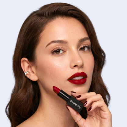 A close-up of a woman with wavy brown hair wearing a deep red lipstick, holding the lipstick tube near her chin. The lipstick tube is black with white text that reads 'JustBaked'.