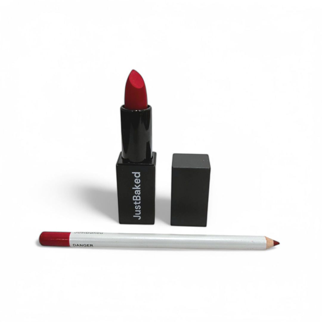 A red lipstick in a black tube with its cap removed, placed next to a white lip liner pencil with a red tip. The lipstick tube is printed with the brand name 'JustBaked'.